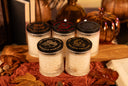 Five small jars of scented candles with black lids are arranged on a wooden board with autumn-themed props including dried leaves, books, pinecones, and decorative pumpkins. The lids show different designs and text including 'The Velvet Noble' and scent names like 'Vanilla Harvest' and 'Golden Twilight'.
