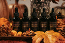 Five black dropper bottles labeled 'VELVET DROPS' arranged on a tray with autumn leaves, pinecones, and pumpkins in a warm, styled setting.