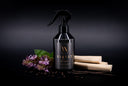 A black trigger spray bottle labeled 'VELVET home' with a gold monogram logo, styled on a dark background with vanilla beans, purple flowers, and black pepper.