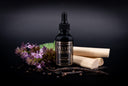 A bottle of 'Velvet Drops' luxury warmer  oil, styled with patchouli flowers, vanilla beans, and wood on a dark background.