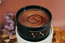 A close-up of a black candle jar filled with a gold swirled wax with glitter, resting on a white ribbed pedestal warmer. The jar has a gold-toned label reading 'VN The Velvet Noble'.
