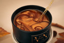 A close-up of a black candle jar filled with a marbled brown liquid, with a gold glittery dropper resting inside. The jar has a black label with gold lettering that reads 'VN The Velvet Noble'.