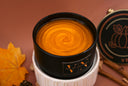A black candle jar filled with a warm amber-orange shimmering wax, displayed in a white ribbed holder with autumn-themed props including a maple leaf and cinnamon sticks on a solid brown background.