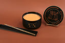 A black candle jar filled with pale orange wax sits on a matching black tray, with the lid displayed on a small stand beside it. The lid and jar are printed with a pumpkin illustration and the words 'VELVET PUMPKIN'; the tray also appears to carry the same branding.