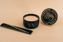 A dark jar candle with a gold-accented label reading 'VN VANILLA HARVEST' next to its matching lid on a stand, with a gold decorative tool in a black tray on a warm beige background.