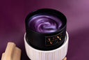 A close-up of a wickless candle with a purple shimmering marbled wax surface, resting in a white ribbed ceramic warmer against a deep purple background. A small gold label on the tin reads 'VN THE VELVET NOBLE'.