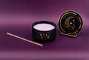 A black jar candle with pale purple wax sits open on a purple background, alongside a gold decorative wand and a round black-and-gold lid labeled 'Golden Twilight.'.