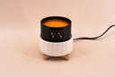A black cylindrical candle vessel with a gold logo sits inside a white ribbed ceramic warmer with small wooden feet, on a warm beige background. A black power cord extends to the right.
