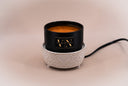 A black candle jar with amber wax sits inside a white textured ceramic warmer base on a warm beige background. The jar features a gold square label with the brand name 'VN' and the phrase 'The Velvet Noble'.