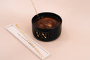 A black candle jar with a gold logo, filled with a brown and gold marbled wax, shown next to its white rectangular tray  on a solid pale tan background.