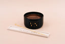 A black jar candle with a gold and black logo, displayed next to a white resting tray on a solid pale tan background.