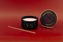 A black candle jar with wax sits open on a red studio background, alongside its matching lid on a small stand. The lid features a gold circular border and a red apple-and-canopy graphic with the words 'NOBLE APPLE.