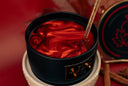 A close-up of a black metal candle jar filled with a glossy, marbled red wax, with a luster wand inserted into the surface. The tin has a gold logo plate on the front and sits on a white ceramic warmer.
