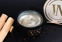 A black jar candle with a lid featuring gold lettering, shown open with a swirled white wax surface. The candle is styled on a dark background with wooden sticks, scattered peppercorns, and part of the branded lid visible.