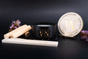 A luxury wickless candle from The Velvet Noble, featuring a matte black jar candle with a gold logo, its matching white lid on a small stand, a white wooden wick set, and wooden wicks, styled with dried flowers on a dark background.
