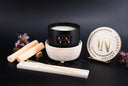 A luxury wickless candle by The Velvet Noble, featuring a white, shimmering wax candle on a textured white ceramic warmer, a round gold-and-white branded tin, and a white box with gold lettering, styled with wooden sticks, black pepper, and purple flowers on a dark background.