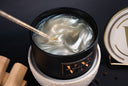 A black candle jar filled with swirling white wax, shown open on a textured white ceramic warmer. A gold-toned clear stirring wand rests inside the tin, and part of a branded white lid with a gold letter V is visible on the right against a dark background.