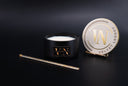 A black candle jar with a gold and black logo plaque, shown next to its round white lid on a small stand and a gold glittery wick, on a dark background. The lid features the name 'VELVET LEGACY'.