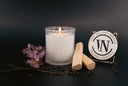 A lit white candle in a textured clear glass jar, styled with two wooden sticks, dark peppercorns, and a small purple flower sprig on a dark background. A round sign reads 'THE VELVET NOBLE VELVET LEGACY' with a monogram.