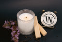 A lit white candle in a textured clear glass jar, styled with two wooden sticks, scattered dark peppercorns, and purple flowers on a dark background. A round sign reading 'THE VELVET NOBLE VELVET LEGACY' with a large 'VN' is visible behind the candle.