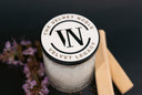 A clear glass candle jar, topped with a white lid printed with the circular 'The Velvet Noble Velvet Legacy' logo. The jar is styled on a dark background with purple flowers and two wooden sticks.