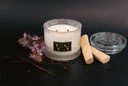 A double-wick scented candle in a textured clear glass jar with a black and gold label reading 'VN', styled on a dark surface with patchouli flowers, vanilla pods, and wooden sticks.