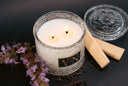 A double-wick scented candle in an ornate clear glass jar with white wax, styled with two wooden wicks, a matching glass lid, dried flowers, and peppercorns on a dark background.