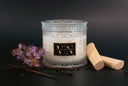 A glass jar candle with white wax and an ornate patterned design, featuring a black label with gold lettering that reads 'VIN THE VELVET NOBLE'. The candle is styled on a dark surface with dried flowers, vanilla beans, and wooden sticks against a dark background.