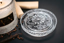 A clear glass decorative candle lid with an embossed floral pattern, shown on a dark surface beside a lit scented candle, peppercorns, and a cinnamon stick.