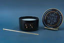 A black candle jar with a gold logo plaque sits next to its matching lid on a small gold stand. The lid features a blue and gold illustration of an umbrella with raindrops and autumn leaves, with the words 'VN' and 'AUTUMN RAIN'.