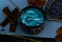 A black candle jar with a swirled blue and teal wax design, shown on a blue surface with dried leaves, cinnamon sticks, and a gold glitter candlestick. Part of the lid with an umbrella illustration is visible on the right.