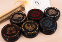 Six round black cosmetic tins with lettering and illustrated lids, arranged on a light beige surface. A white drawstring pouch and a rectangular box are partially visible in the background.
