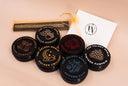 Six round black candle tins with illustrated lids arranged on a beige surface, alongside a white branded card and a small gold drawstring pouch. The candles are labeled with names such as Velvet Pumpkin, Vanilla Harvest, Golden Twilight, Autumn Rain, Noble Apple, and Vintage Pumpkin.