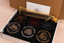 Six round black candle tins with designs and text, displayed in a white gift box with black filler, alongside a gold sheer pouch and a branded card.