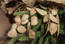Close-up of a holiday tree featuring textured beige fabric leaves and green pine branches, with a wicker bell edge visible at the top.