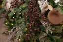 Close-up of a decorated Christmas tree branch with green pine needles, brown glittery boxwood branch sprays, warm white fairy lights, a cream ribbon, and a round brown ornament.