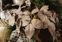 Close-up of a decorative artificial leaf arrangement in warm taupe and beige tones with glittery texture, photographed in a dark festive setting with soft holiday lights.