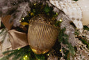 A close-up of a ribbed metallic gold acorn-shaped Christmas ornament nestled among green pine branches, white ribbon, and warm string lights.