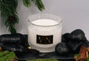 Candle in a decorative glass holder with a black label, surrounded by stones and greenery.