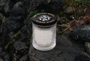 Clear glass candle in a decorative container with a black lid on a stone surface.