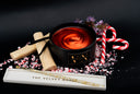A black candle tin with a red-orange swirled wax fill sits on a black surface, surrounded by candy cane pieces, pink peppercorn, two wooden wick props, a gold scented wax dip pen, and a white tray labeled 'THE VELVET NOBLE'. The candle tin has a visible 'VN' label.