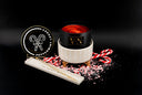 A black jar candle with a cream ribbed base sits on a black background, surrounded by candy cane props, crushed candy cane pieces, and a branded lid leaning beside it. The lid reads 'Peppermint Legacy'