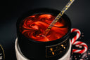 A close-up of a black candle vessel filled with a glossy marbled red wax, with a clear glittery spoon resting inside. The candle sits on a white base against a dark background, with candy canes visible in the blurred background.