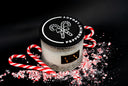 A glass jar candle with a black lid featuring a white candy cane graphic and the text 'PEPPERMINT LEGACY', styled with red-and-white candy canes and crushed candy cane pieces on a black background.