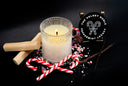 A lit candle in a textured clear glass holder sits on a black surface with candy cane pieces, wooden sticks, a vanilla bean, and a round black label reading 'The Velvet Noble Peppermint Legacy' behind it.