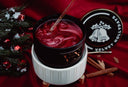 A festive wickless candle in a black jar with a white ribbed warmer, filled with dark red wax and a glass thermometer, set on a red fabric background with cinnamon sticks, pine branches, and the branded lid nearby.