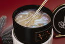 A close-up of a black candle jar with a gold label, filled with a marbled wax design in cream, gold, and iridescent tones. A gold-and-black wick is visible, and part of the matching black lid sits to the right against a red background.
