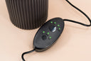 A close-up of a black oval electronic control module with green LED indicator lights and two round buttons, connected by a black cable, set against a warm beige background with a dark ribbed cylindrical object partially visible behind it.