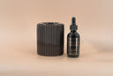 A dark brown ribbed cylindrical fragrance warmer next to a small black dropper bottle labeled 'VELVET DROPS' on a warm beige background.