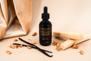 Black bottle of 'Velvet Drops' with vanilla beans and a wood on a beige background